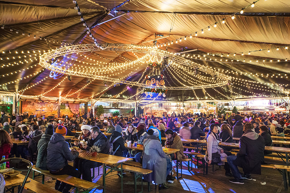 Inside a large tent in a winter wonderland setting, fully adorned with lights, there are numerous tables at full capacity.