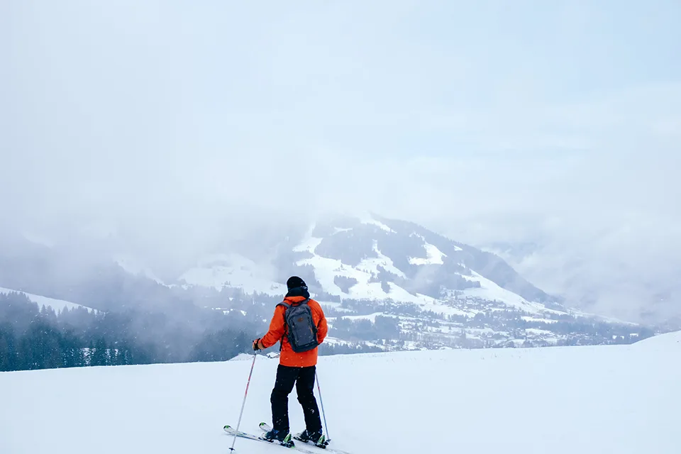 Man on skis at the top of a snowy hill, wearing an orange winter coat, looking towards distant snow-covered mountains