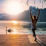A woman practicing yoga by the lake, saluting the Sun with a swan passing by
